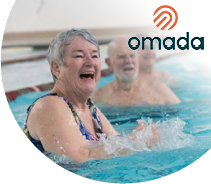 Three older Alaskans have fun taking a water aerobics class in the pool at a YMCA in Anchorage, AK. A woman smiles and splashes in front and two men exercise behind her.