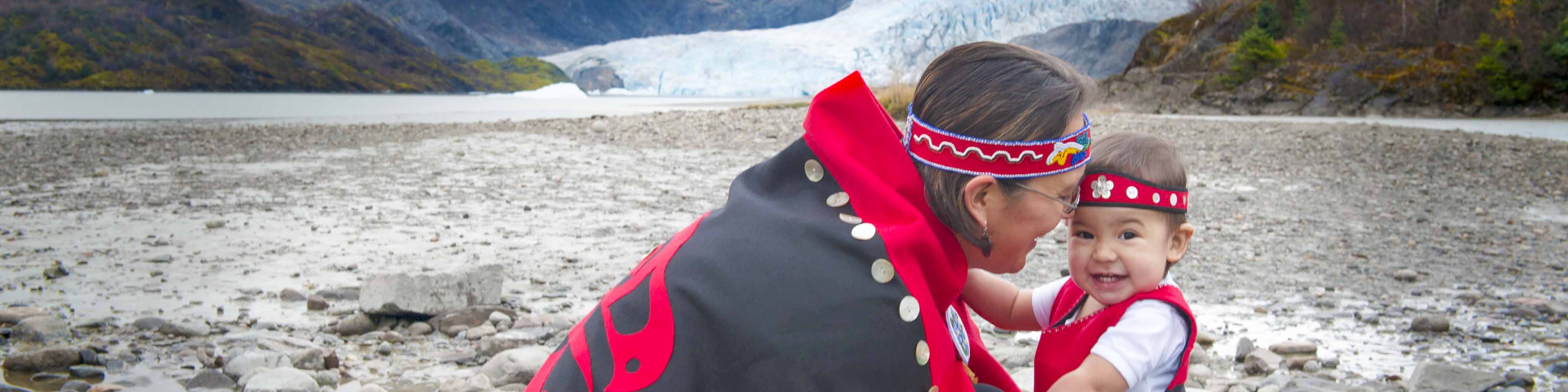 A woman in regalia kneels down face-to-face with a child at the Mendenhall Glacier